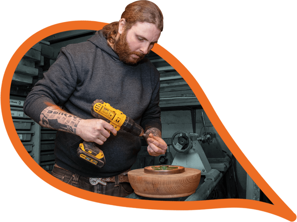 A social entrepreneur with long red hair and a beard uses a drill in a wood workshop. 