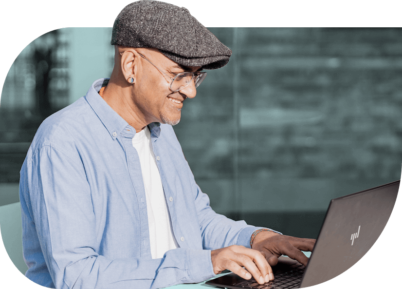 Sonny Thaker, a social entrepreneur wearing a blue shirt, grey flat cap and rainbow striped stud earring smiles, typing on a laptop.