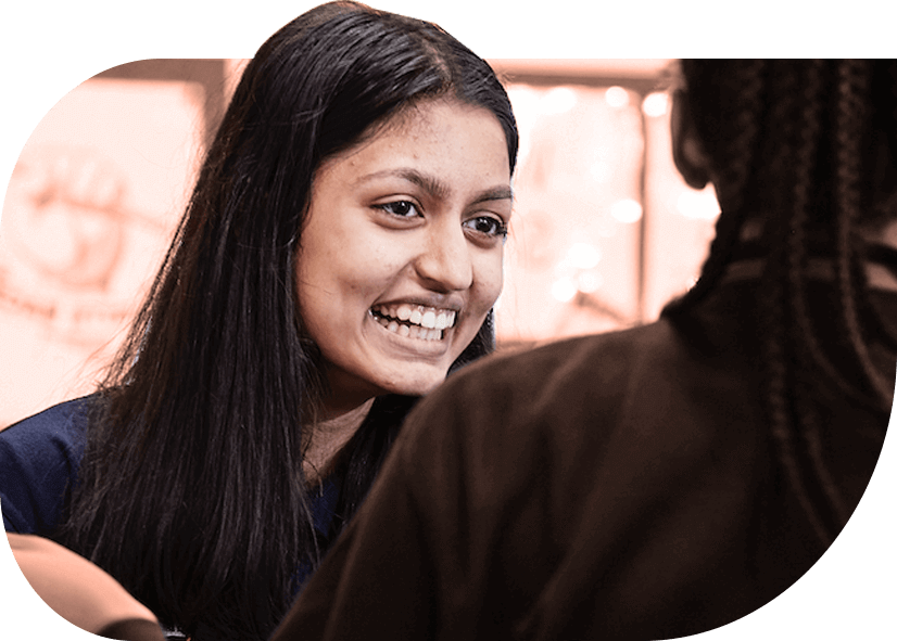 A young person with long straight dark brown hair smiles in a workshop.