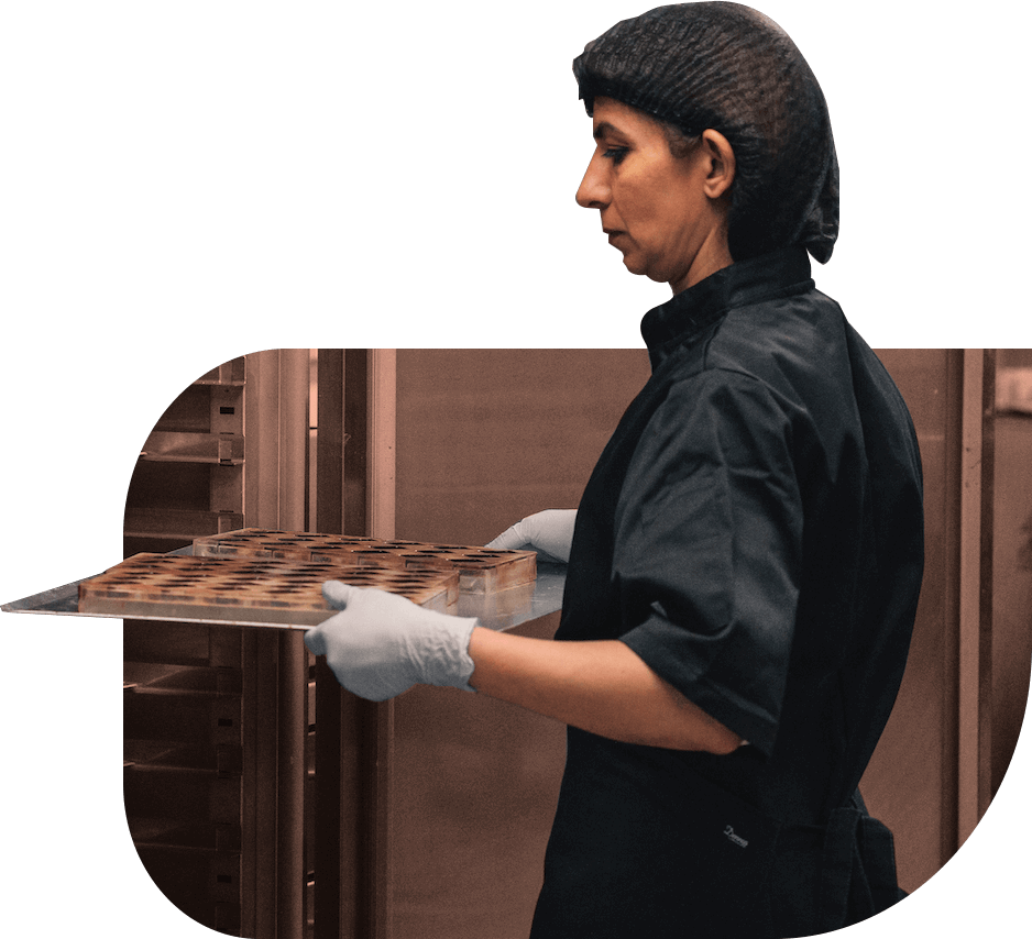 A social entrepreneur lifts a tray of chocolates in a commercial kitchen. She wears gloves, a black chef jacket and a hairnet.
