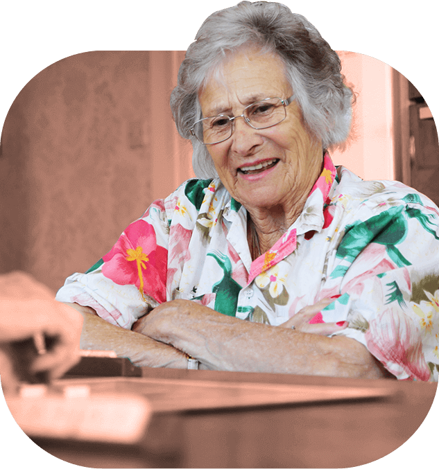 An older person with short grey hair wearing a floral shirt and glasses smiles, looking at a piece of paper on the table.