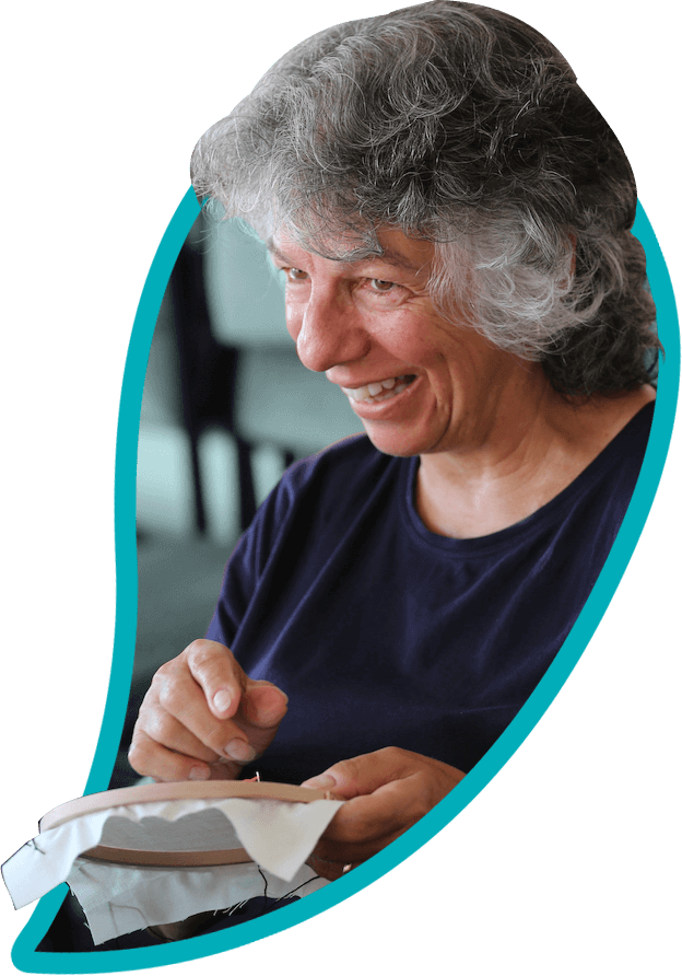 An older person with short grey wavy hair smiles, using an embroidery hoop.