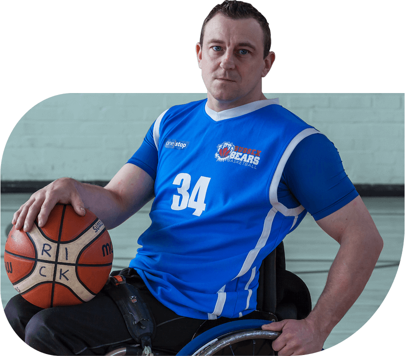 Ricky Perrin, a social entrepreneur using a wheelchair sits wearing blue sportswear holding a basketball, with a serious facial expression.