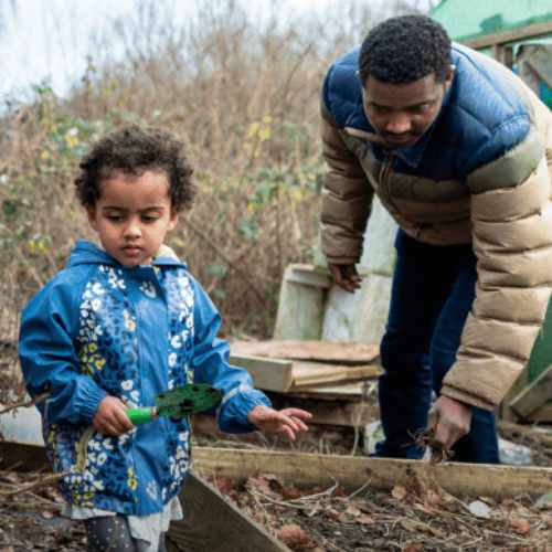 A child in a blue raincoat holds a mini trowel in a community garden.